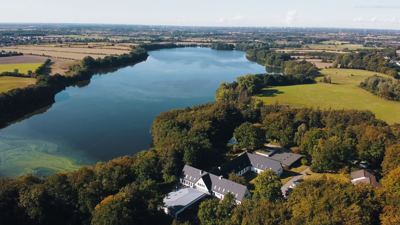 Luftaufnahme der Akademie Sankelmark: Ein weißer Gebäudekomplex mit dunklen Dächern liegt eingebettet in dichten Wald direkt am Ufer eines großen, blauen Sees. Im Hintergrund erstreckt sich eine weite Landschaft mit Feldern und Windrädern am Horizont.